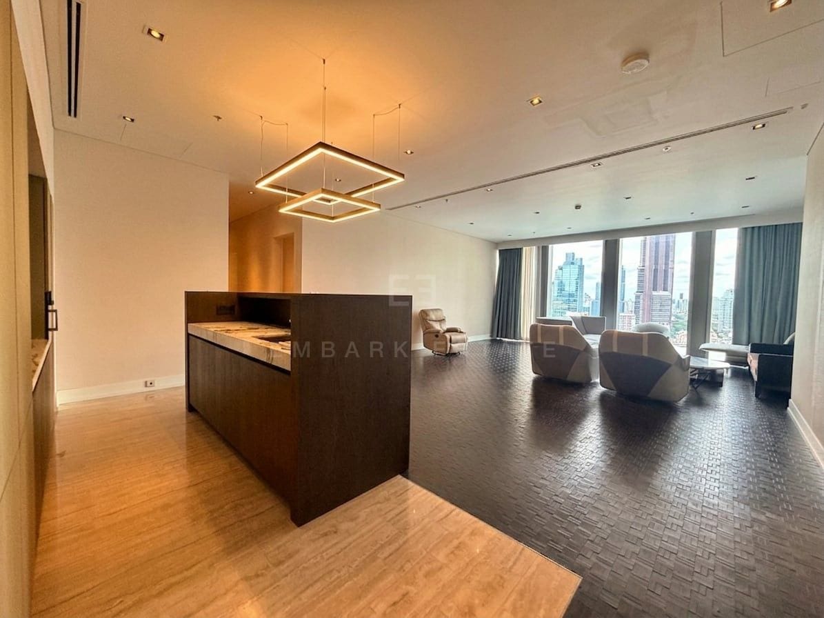 The living and dining area of a three-bedroom residence at The Ritz-Carlton Residences Bangkok seen past the kitchen island, with square ring pendants above, a travertine entrance floor and seating grouped at the windows