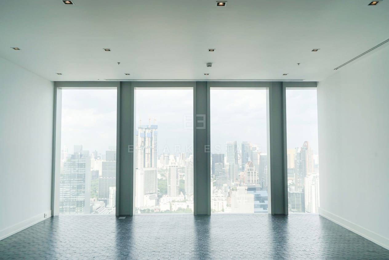 The empty living room of a three-bedroom residence at The Ritz-Carlton Residences Bangkok, with full-height windows across one wall looking over the Sathorn and Silom skyline, and dark herringbone parquet