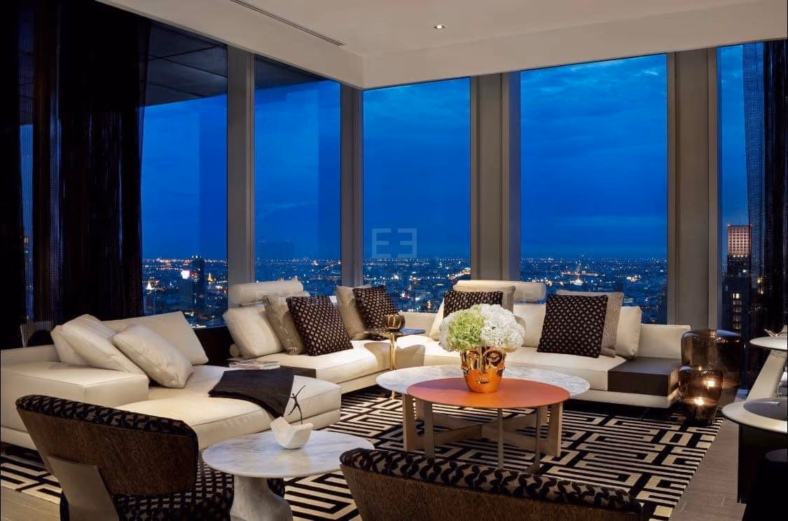 The living room of a two-bedroom residence at The Ritz-Carlton Residences Bangkok at dusk, pale sofas on a patterned rug with the city through full-height glass