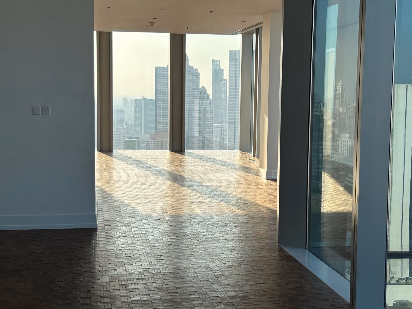The empty living room of a two-bedroom residence at The Ritz-Carlton Residences Bangkok, low sun across bare parquet and full-height glass onto the city