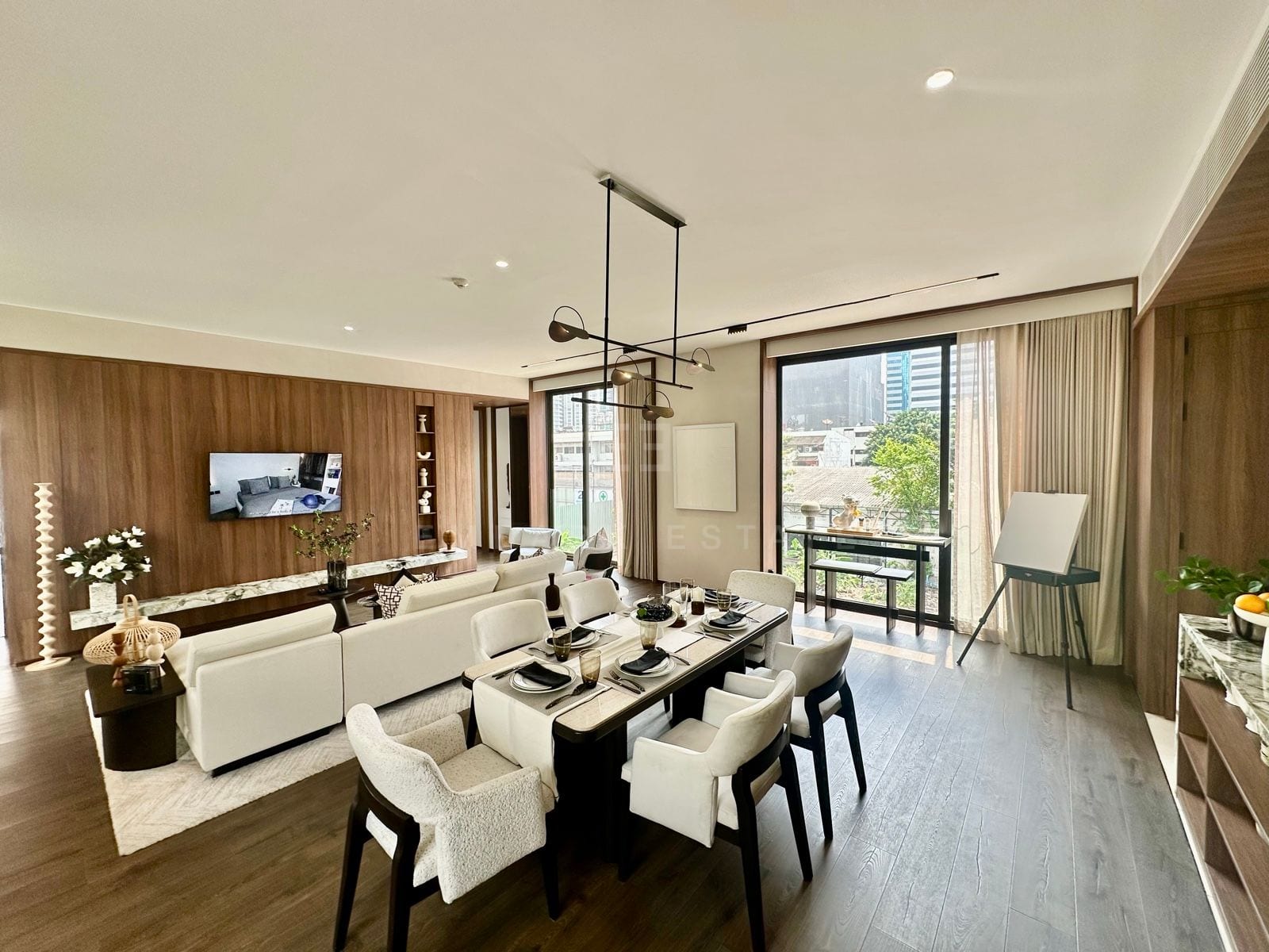 Open-plan living and dining room with a white sectional sofa, six-seat dining table, wood-panelled feature wall with wall-mounted television and a floor-to-ceiling glass door to the balcony, a 2-bedroom villa at ROMM Convent, Bangkok