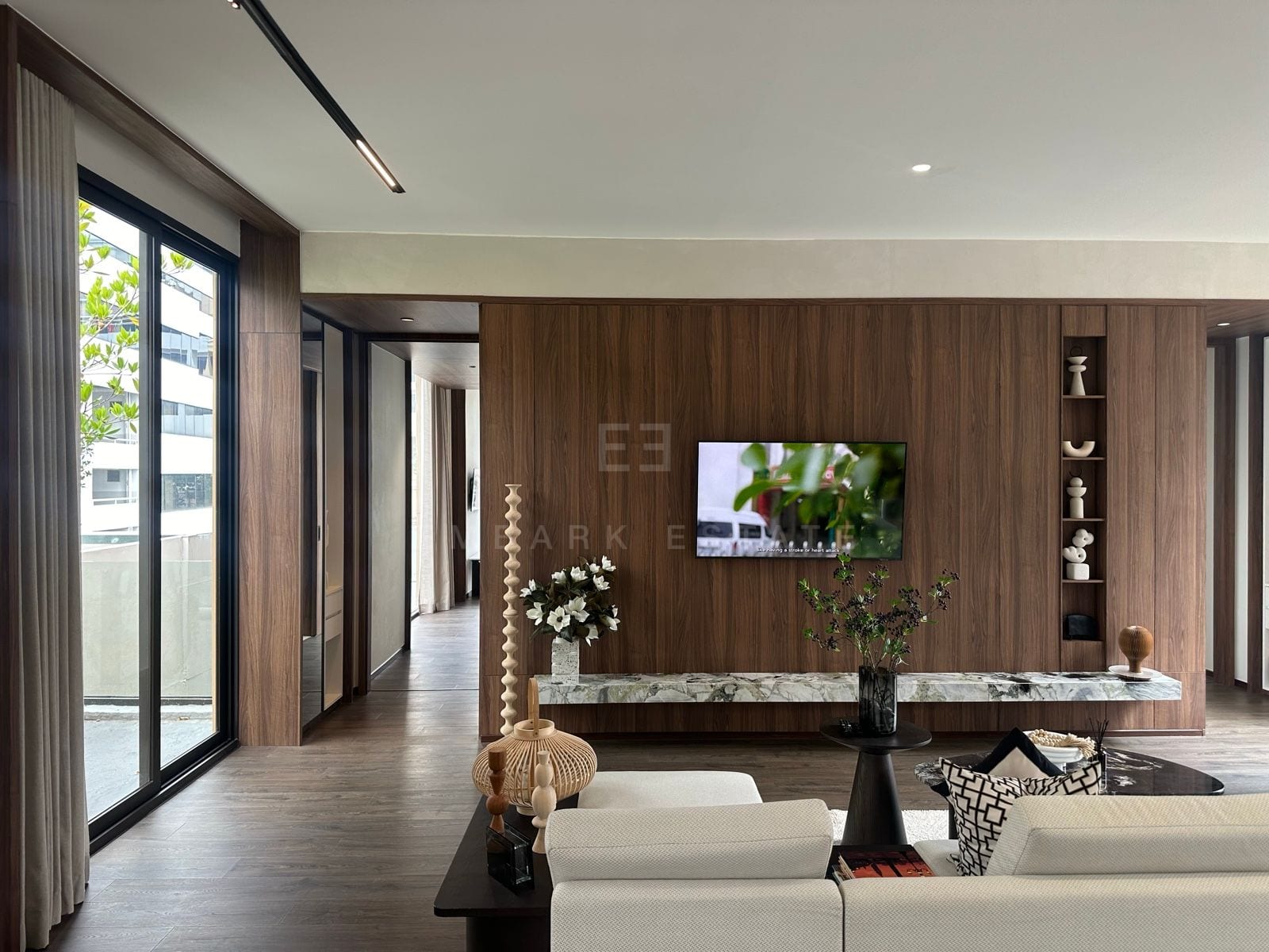 Living room with a walnut feature wall, wall-mounted television above a green marble console, cream sofa and floor-to-ceiling sliding glazing onto an outdoor area with greenery, a 2-bedroom villa at ROMM Convent, Bangkok