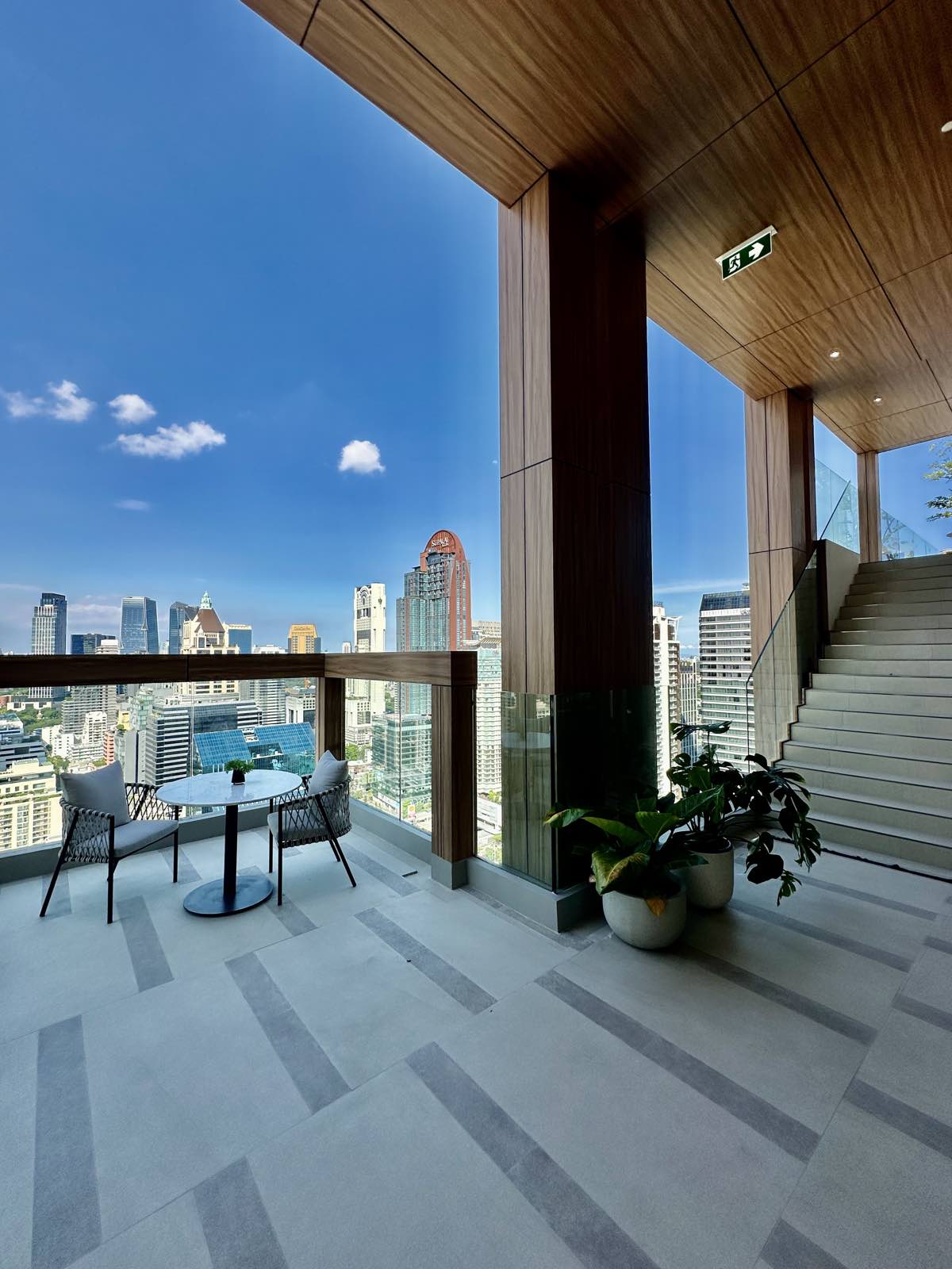 Open-air sky terrace with woven chairs, a round table and planting above the Bangkok skyline at ROMM Convent.