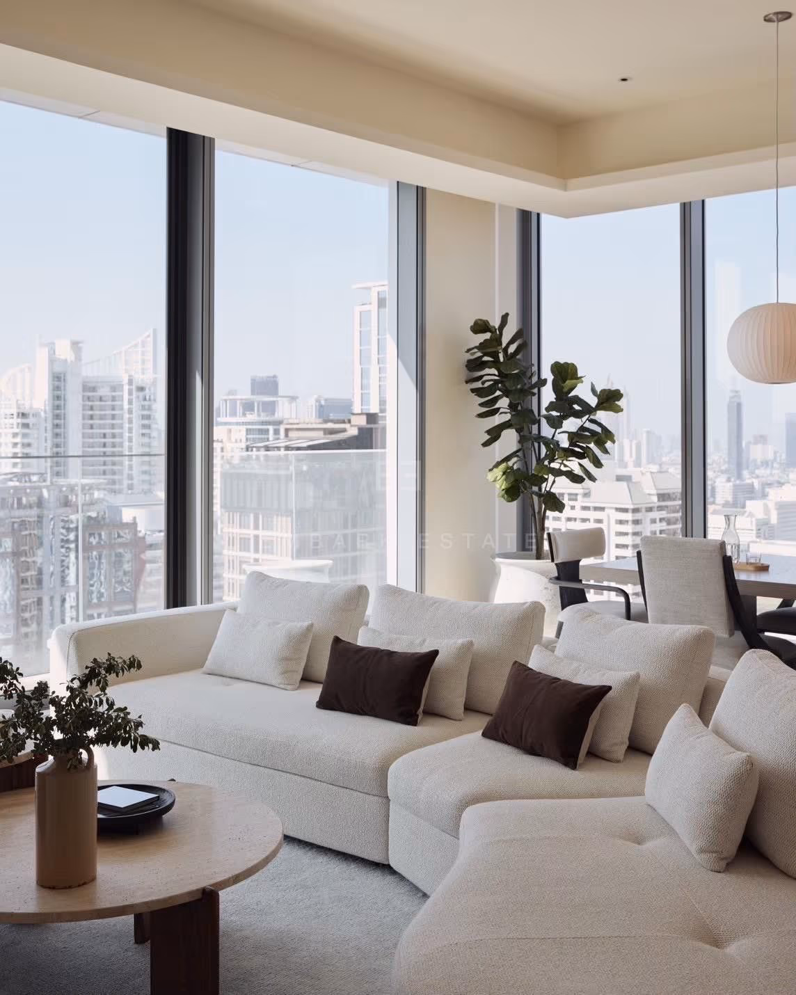 A cream sofa beside the corner windows of a 3-bedroom residence at Scope Langsuan, Bangkok, with the dining table and city towers beyond
