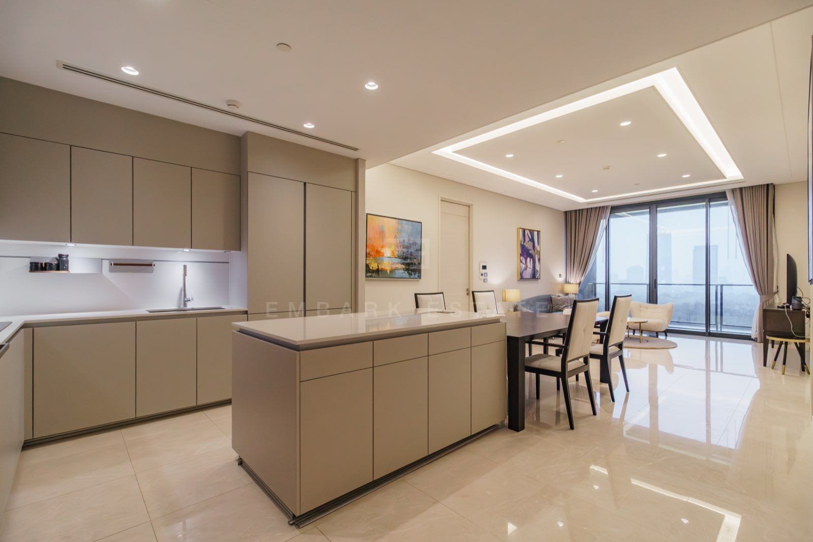 The open kitchen of the two-bedroom residence at the Residences at Sindhorn Kempinski, Bangkok, with a taupe island, a dining table and windows over the central Bangkok skyline