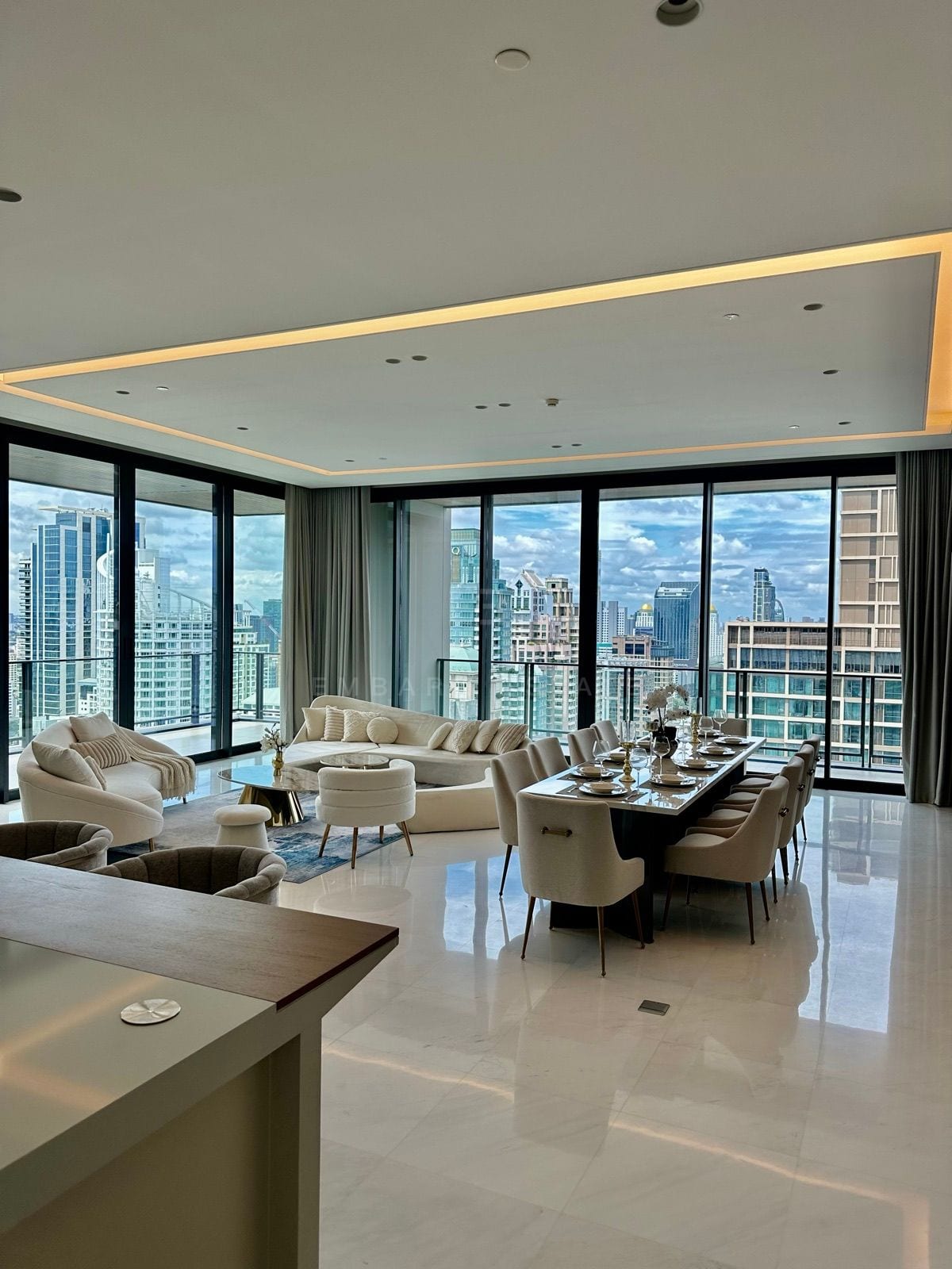 ห้องนั่งเล่นและห้องอาหารของจูเนียร์เพนต์เฮาส์ที่ The Residences at Sindhorn Kempinski Hotel Bangkok พร้อมโซฟาทรงโค้งสีครีมและกระจกสูงจรดเพดานมองเห็นเส้นขอบฟ้า