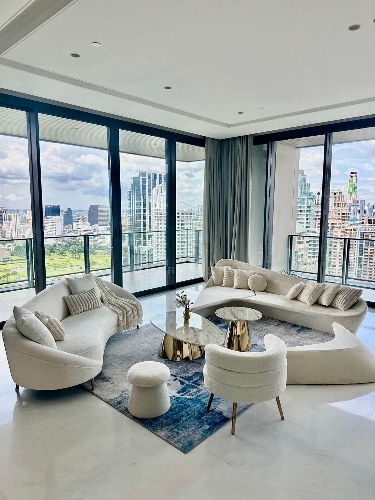ห้องนั่งเล่นของจูเนียร์เพนต์เฮาส์ที่ The Residences at Sindhorn Kempinski Hotel Bangkok พร้อมโซฟาทรงโค้งสีขาวสองตัวและกระจกสูงจรดเพดานมองเห็นเส้นขอบฟ้าใจกลางกรุงเทพฯ