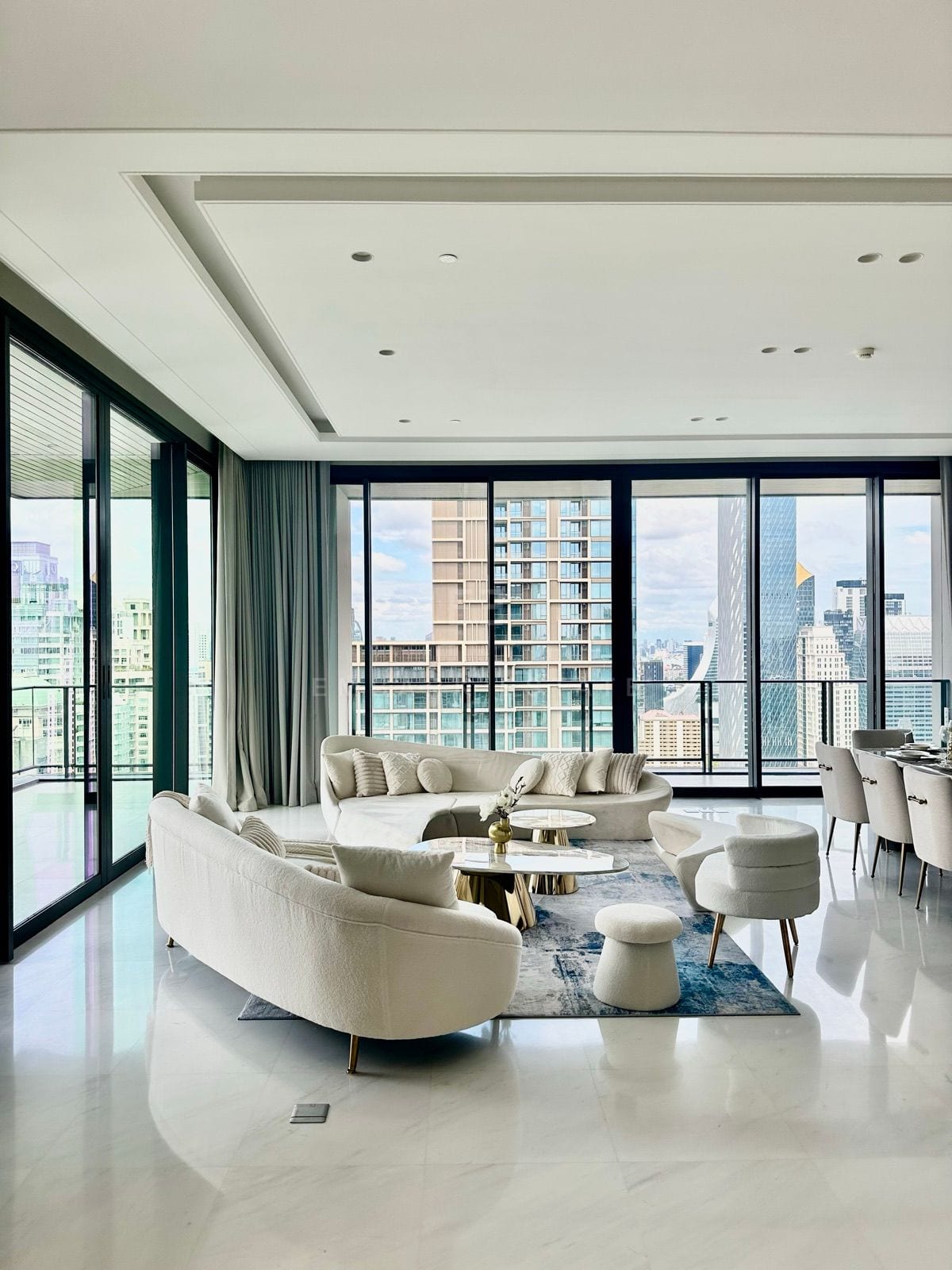 ห้องนั่งเล่นของจูเนียร์เพนต์เฮาส์ที่ The Residences at Sindhorn Kempinski Hotel Bangkok พร้อมโซฟาทรงโค้งสีขาวและกระจกสูงจรดเพดานมองเห็นเส้นขอบฟ้าใจกลางกรุงเทพฯ