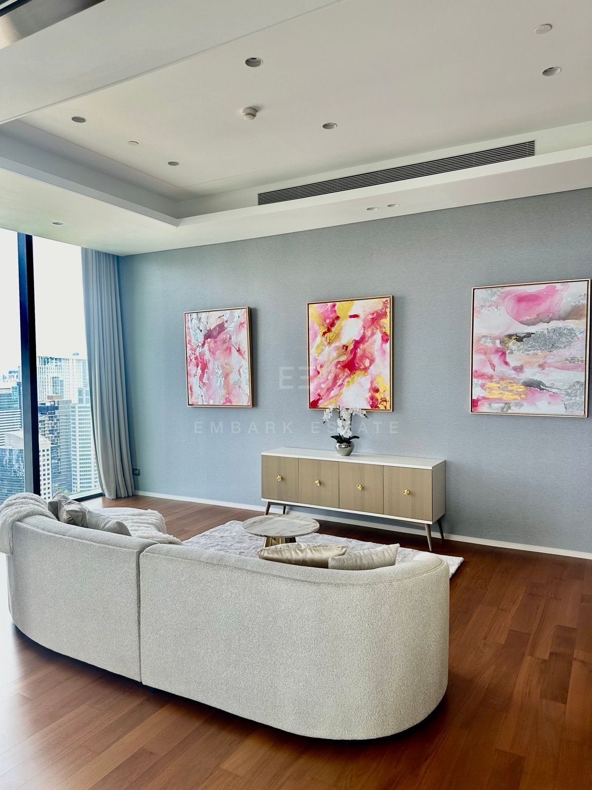 A sitting area of the junior penthouse at the Residences at Sindhorn Kempinski, Bangkok, with a curved sofa, wood floor and three framed pink artworks above a console