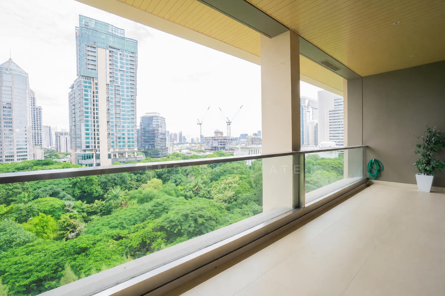 The covered terrace of the two-bedroom penthouse at Sindhorn Residence Langsuan, Bangkok, with a glass balustrade, pale tiled floor, slatted timber ceiling, a potted plant, and treetops and city towers beyond.