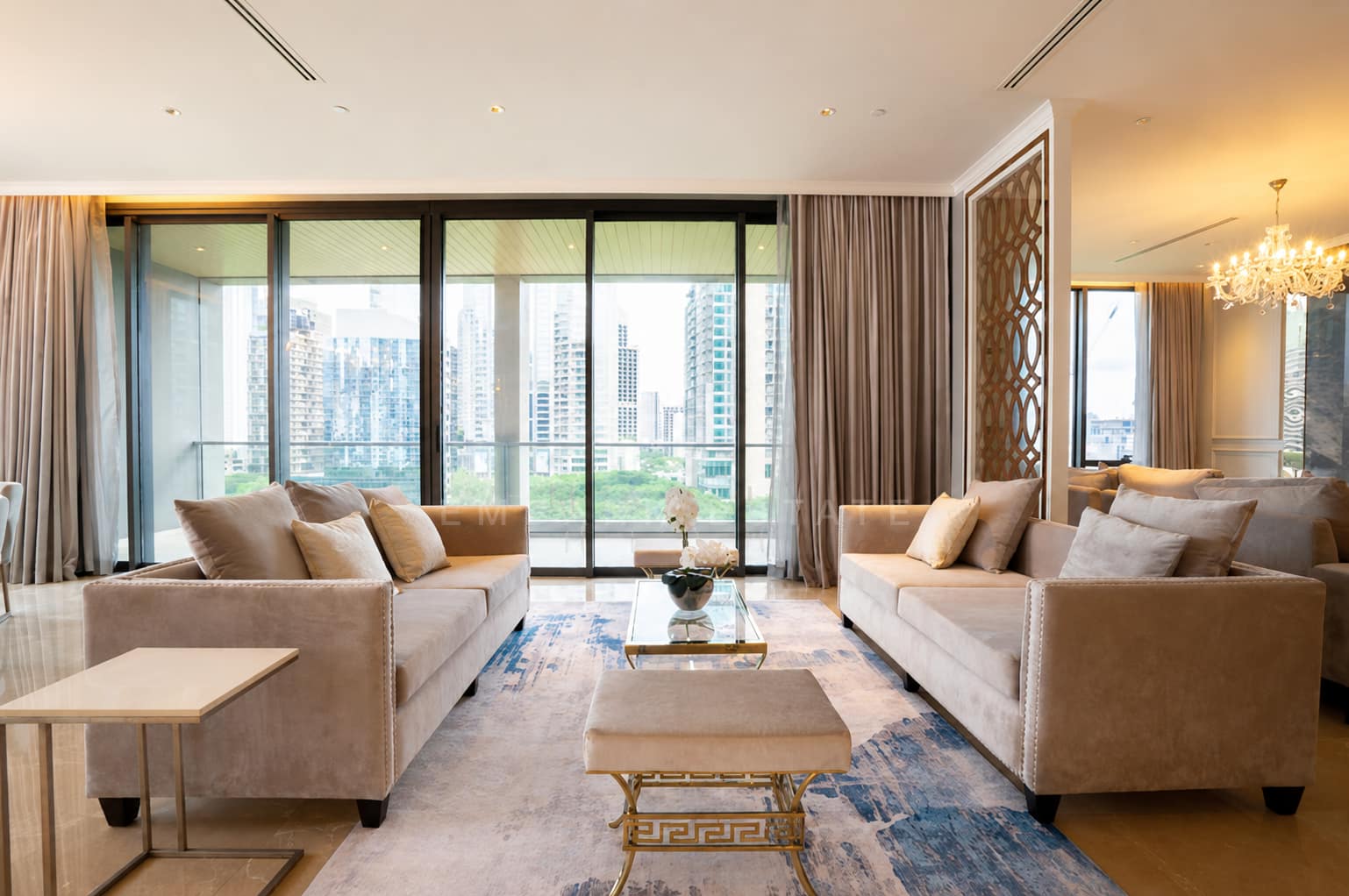 The living room of the two-bedroom penthouse at Sindhorn Residence Langsuan, Bangkok, with two facing beige velvet sofas, a gold and glass coffee table, blue abstract rug, and floor-to-ceiling glass framed by full-length curtains.
