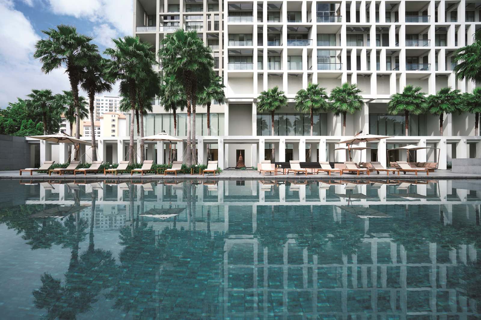 The Sukhothai Residences ลานสระว่ายน้ำในเวลากลางวันพร้อมเตียงอาบแดด ร่ม และต้นปาล์มที่สะท้อนบนผิวน้ำนิ่ง ตัดกับฟาซาดของอาคารพักอาศัย
