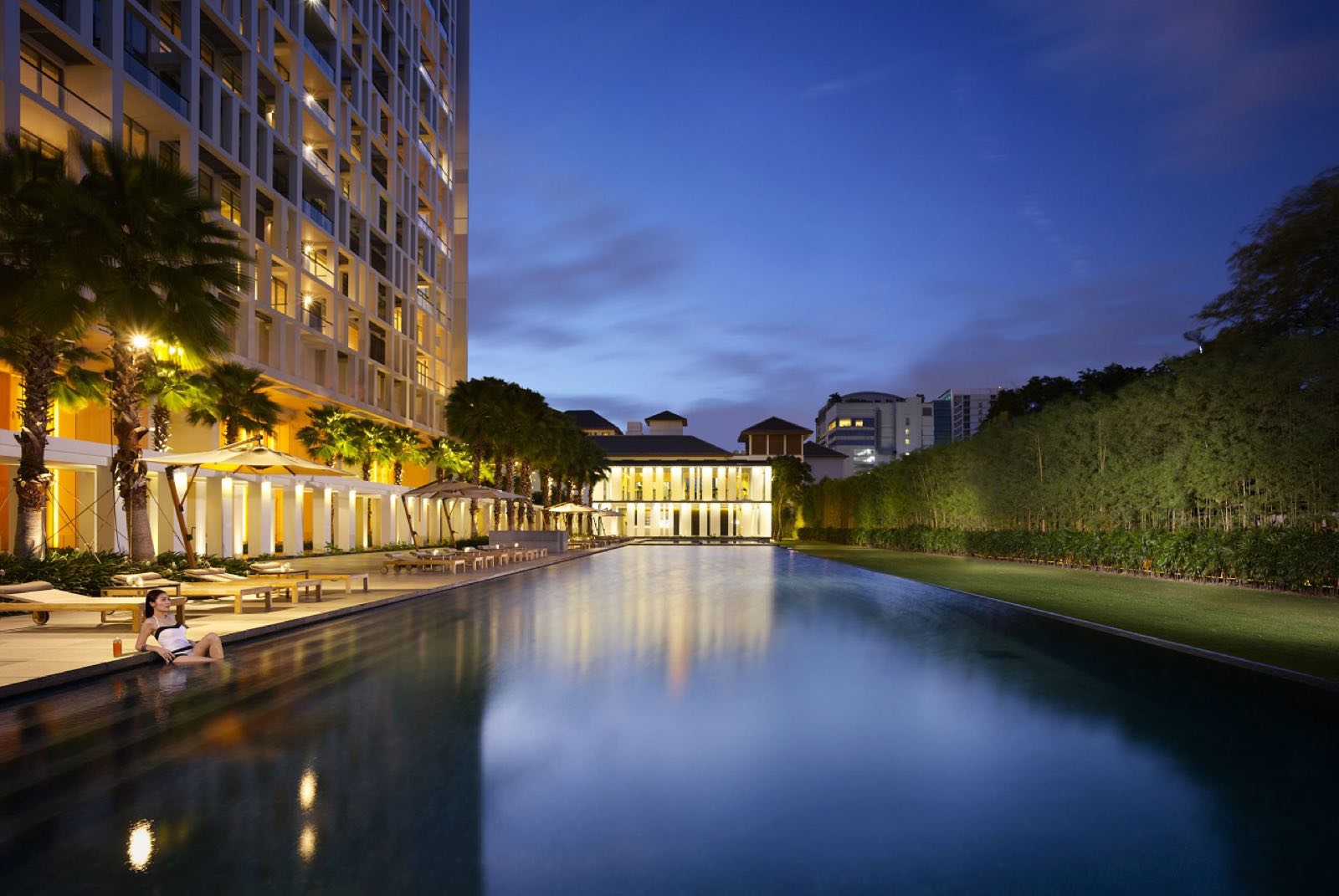 The Sukhothai Residences: Reflecting pool and palm-lined terrace beside the residence tower lit at dusk, with the pavilion behind