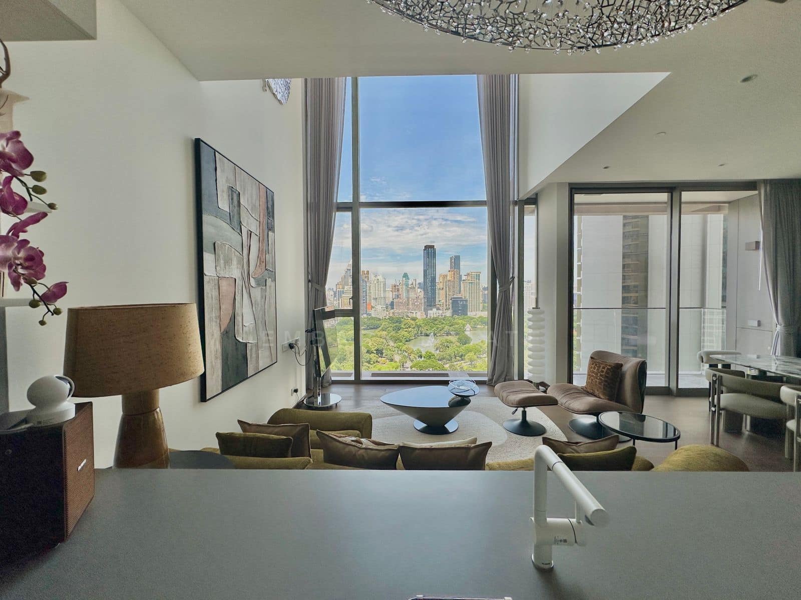 The double-height living room of the duplex at Dusit Parkside, Dusit Central Park, seen from the kitchen counter, a tall window over Lumphini Park, its lake and fountain, and the Sukhumvit skyline