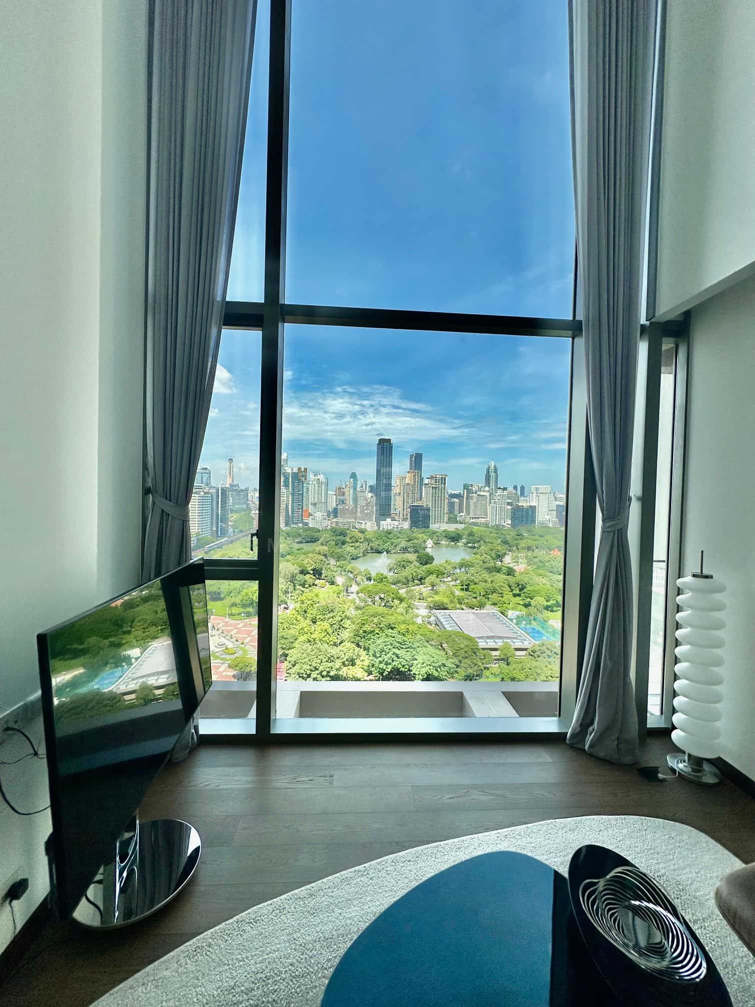 The double-height window of the duplex at Dusit Parkside, Dusit Central Park, framing Lumphini Park, its lake and the towers beyond from the 17th floor