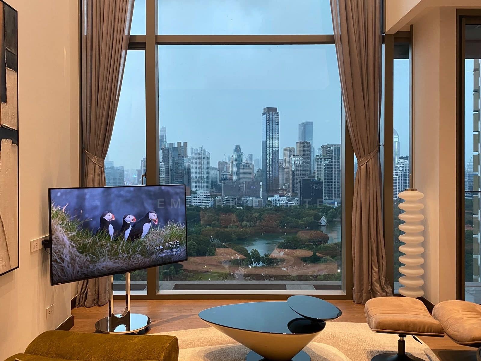 The double-height living room of the duplex at Dusit Parkside, Dusit Central Park, furnished, a full-height window over Lumphini Park and its lake, with a sculpted coffee table and a lounge chair