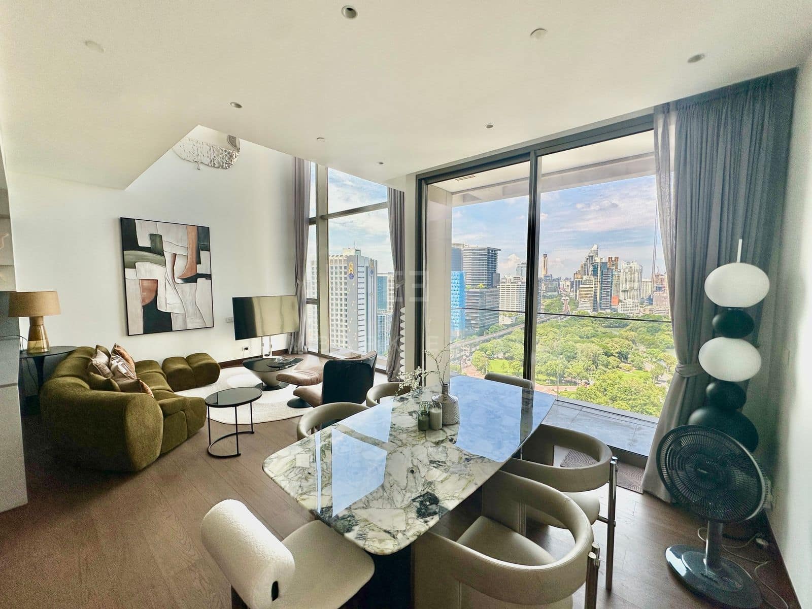 The open living and dining room of the duplex at Dusit Parkside, Dusit Central Park, a green velvet sofa and a marble table beneath a double-height ceiling, the park through full-height glass