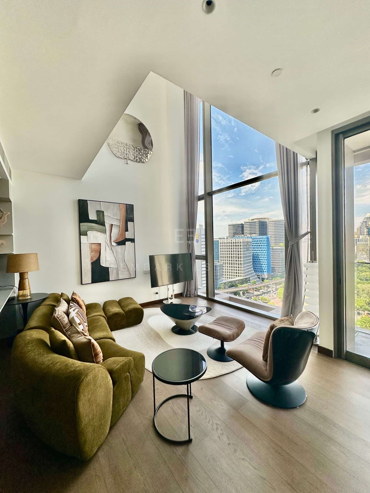 The living room of the duplex at Dusit Parkside, Dusit Central Park, a double-height wall with a sculpted mirror, a curved green sofa and a swivel lounge chair at the tall window