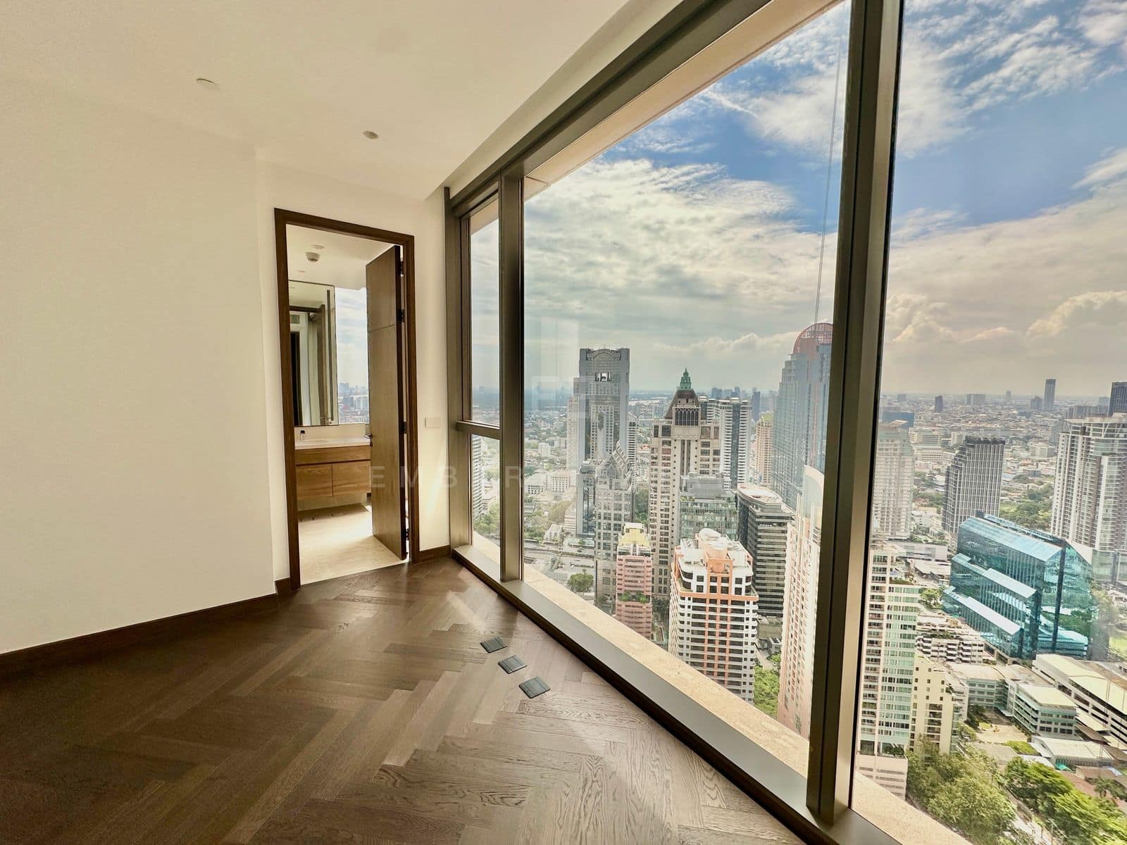 A bedroom of the residence at Dusit Residences, Dusit Central Park, herringbone oak and a full-height corner window over the towers of Silom, with the en-suite bathroom alongside