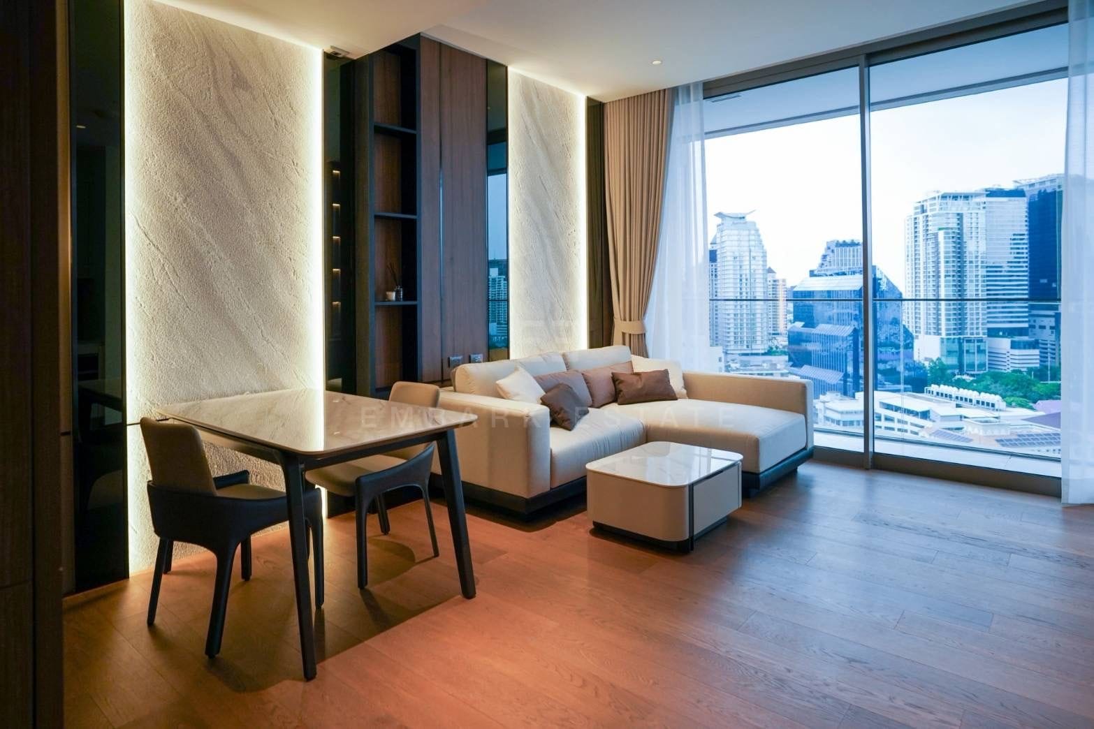 ห้องนั่งเล่นและมุมรับประทานอาหารของยูนิตขนาด 56.6 ตารางเมตร ที่ Dusit Parkside, Dusit Central Park ผนังหินมีไฟส่องด้านหลังโต๊ะอาหารหินอ่อน และโซฟาเข้ามุมสีครีมหน้ากระจกสูงจรดเพดานที่มองเห็นเส้นขอบฟ้าย่าน Silom ยามพลบค่ำ