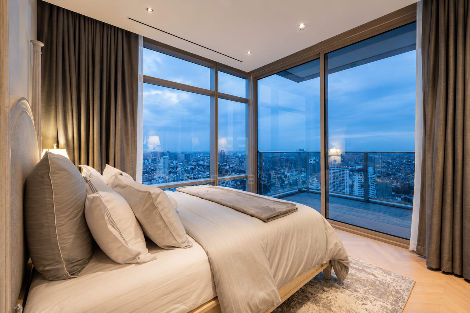 A bedroom at Four Seasons Private Residences, a tall padded headboard beside corner glazing over the river at dusk