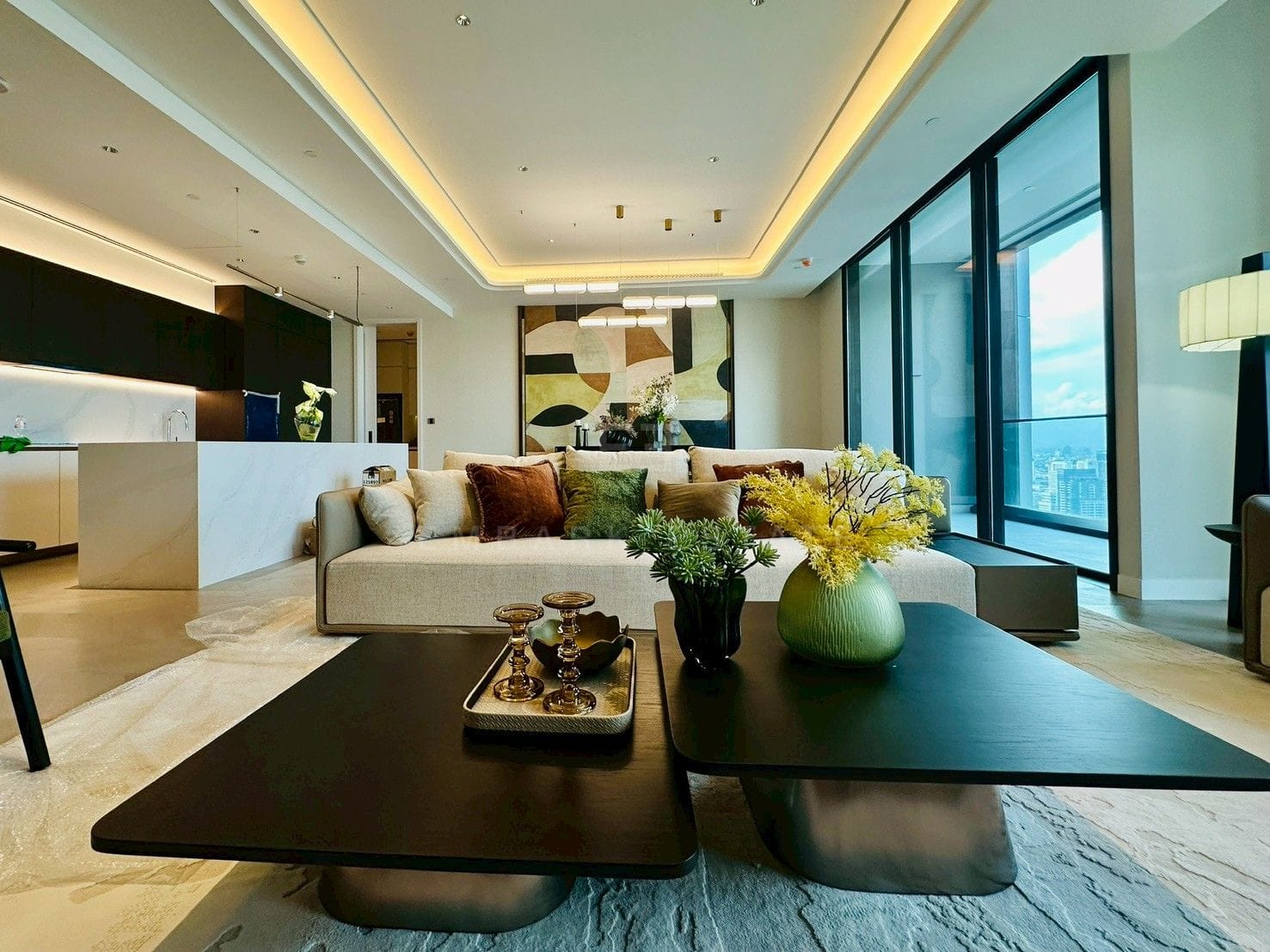 Living room at ONE89 Wireless, Bangkok, with a long linen sofa, twin bronze-based coffee tables and a painted panel wall, the kitchen island beyond
