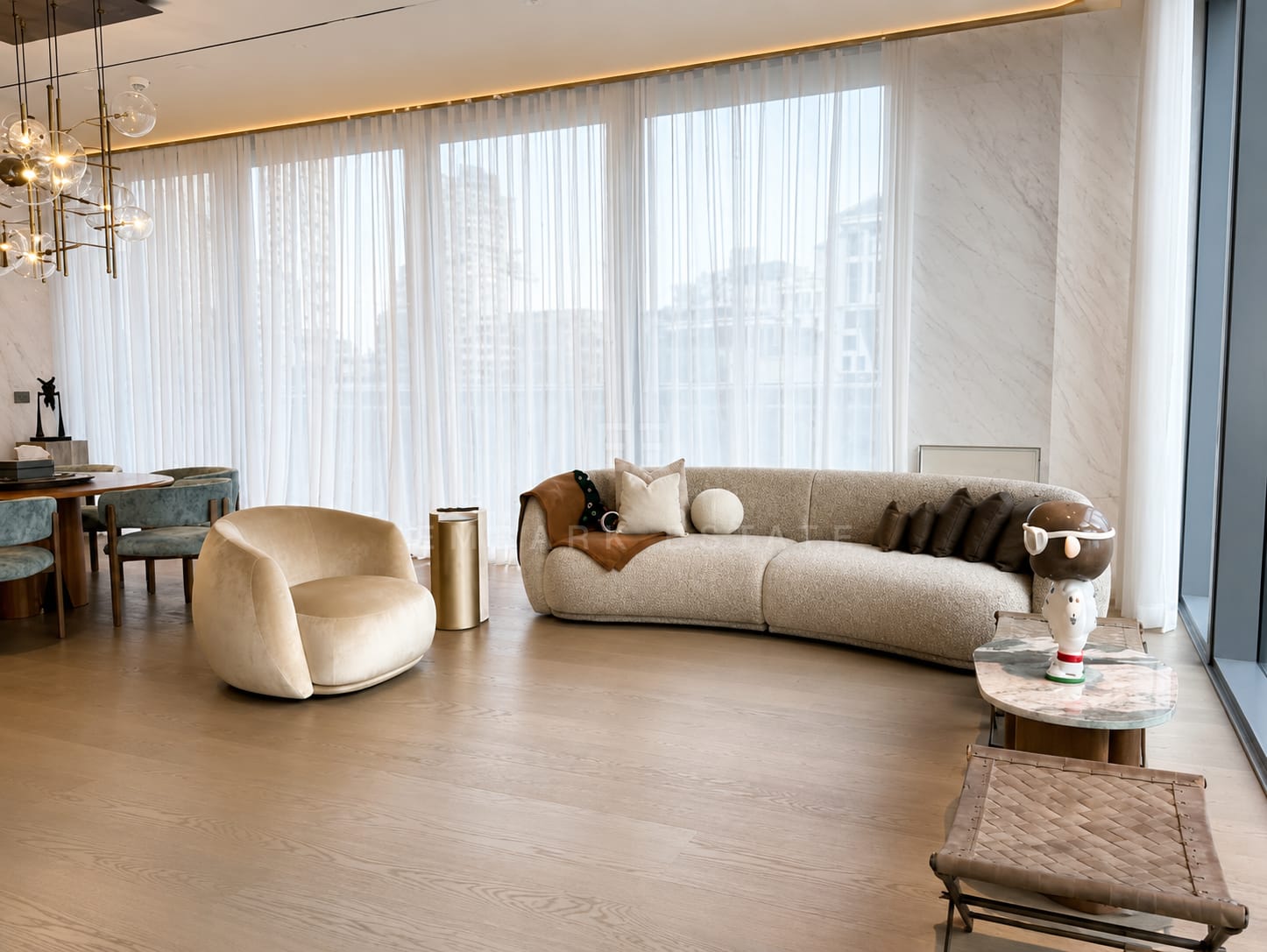 The living room at Scope Langsuan, a curved sofa and bouclé armchair on timber with sheer curtains along the glazing and the dining table beyond