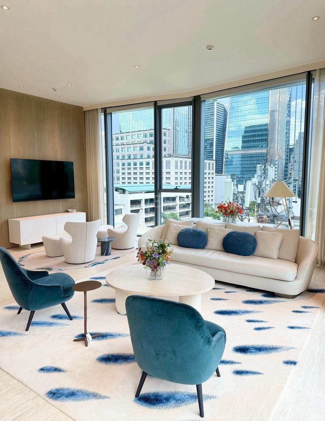 Living room at Scope Langsuan, Bangkok, with a cream sofa, teal velvet armchairs and corner glazing over the Langsuan skyline