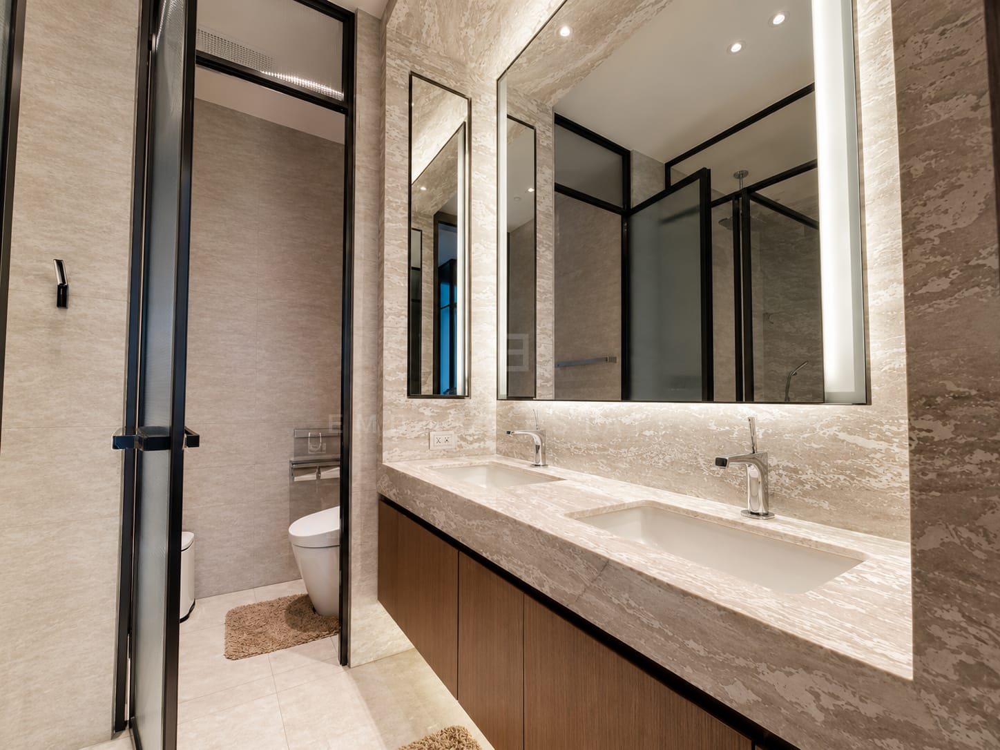 The bathroom at Scope Langsuan, twin basins on a travertine vanity under a backlit mirror with a separate glazed WC