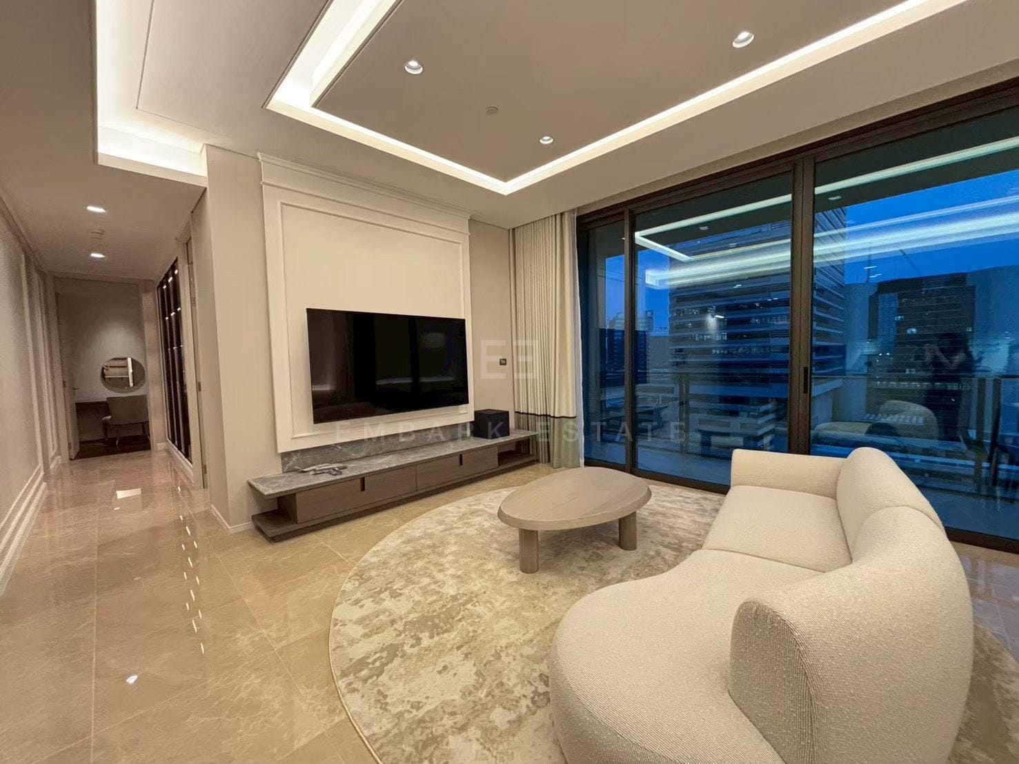 The living room of the 157 square metre residence at Sindhorn Kempinski, a curved bouclé armchair and a round stone table on a marble floor, sliding glass onto the balcony at dusk
