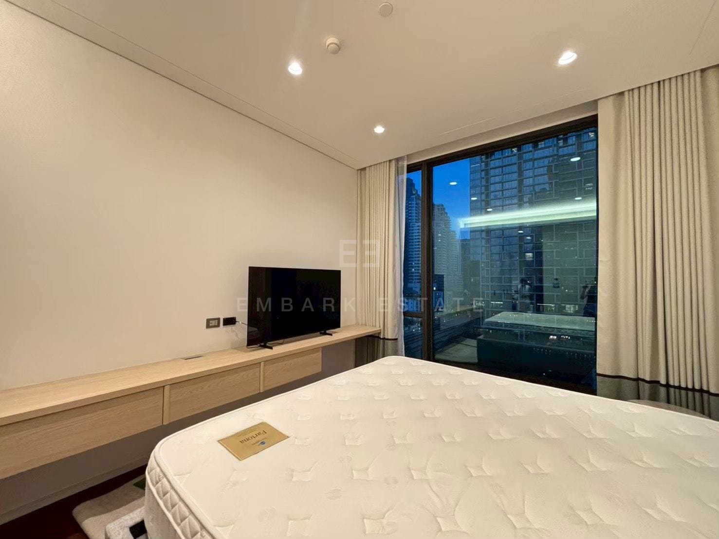 A bedroom at Sindhorn Kempinski, a long timber console beneath the television and glazing onto the balcony at dusk
