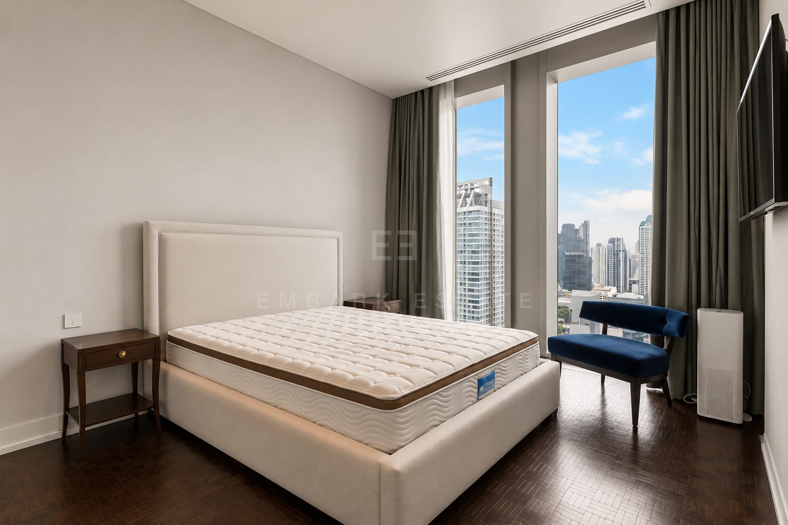 A bedroom at The Ritz-Carlton Residences Bangkok, a panelled headboard and a blue velvet chair beside full-height glazing over the city