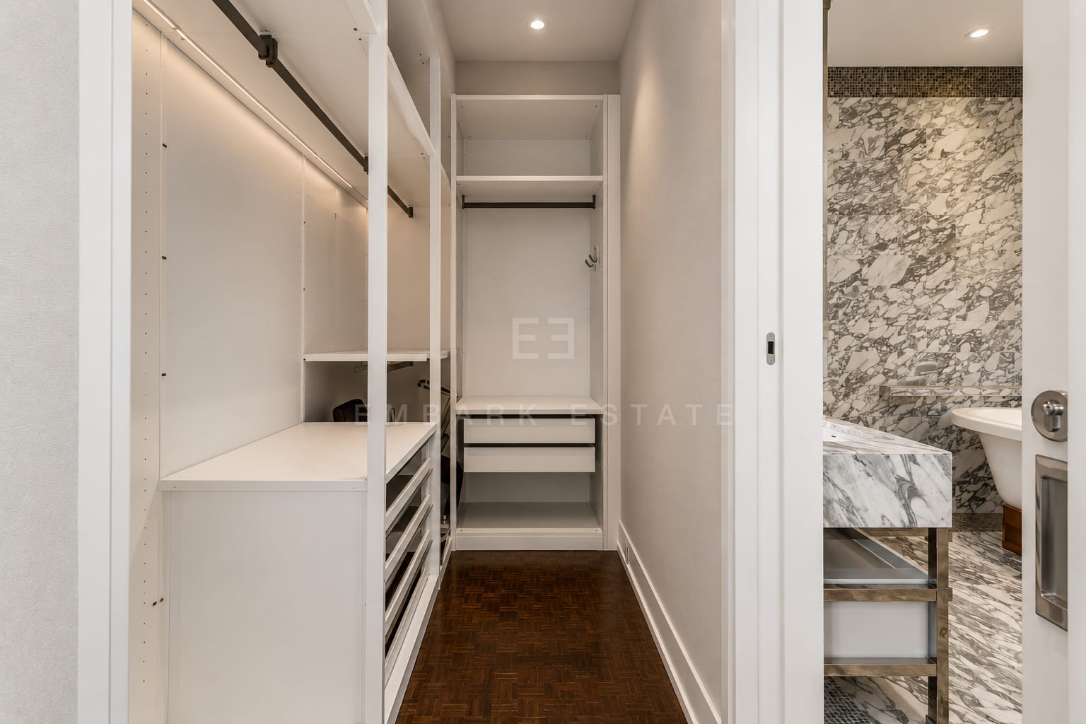 The walk-in wardrobe at The Ritz-Carlton Residences Bangkok, fitted rails and drawers in white with the marble bathroom through the door