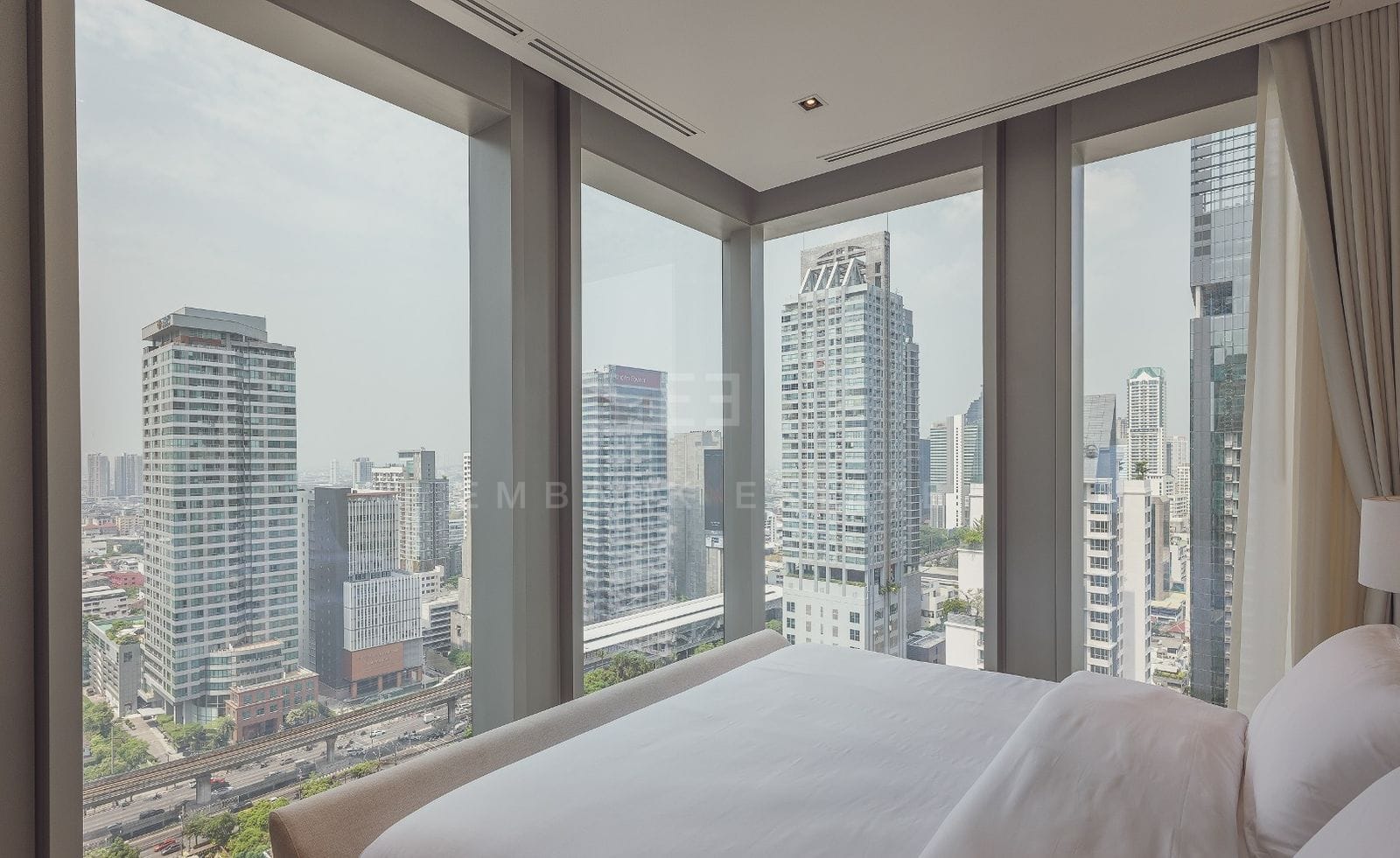 A bedroom at The Ritz-Carlton Residences Bangkok, the bed set against corner floor-to-ceiling glass with the towers and the BTS line below