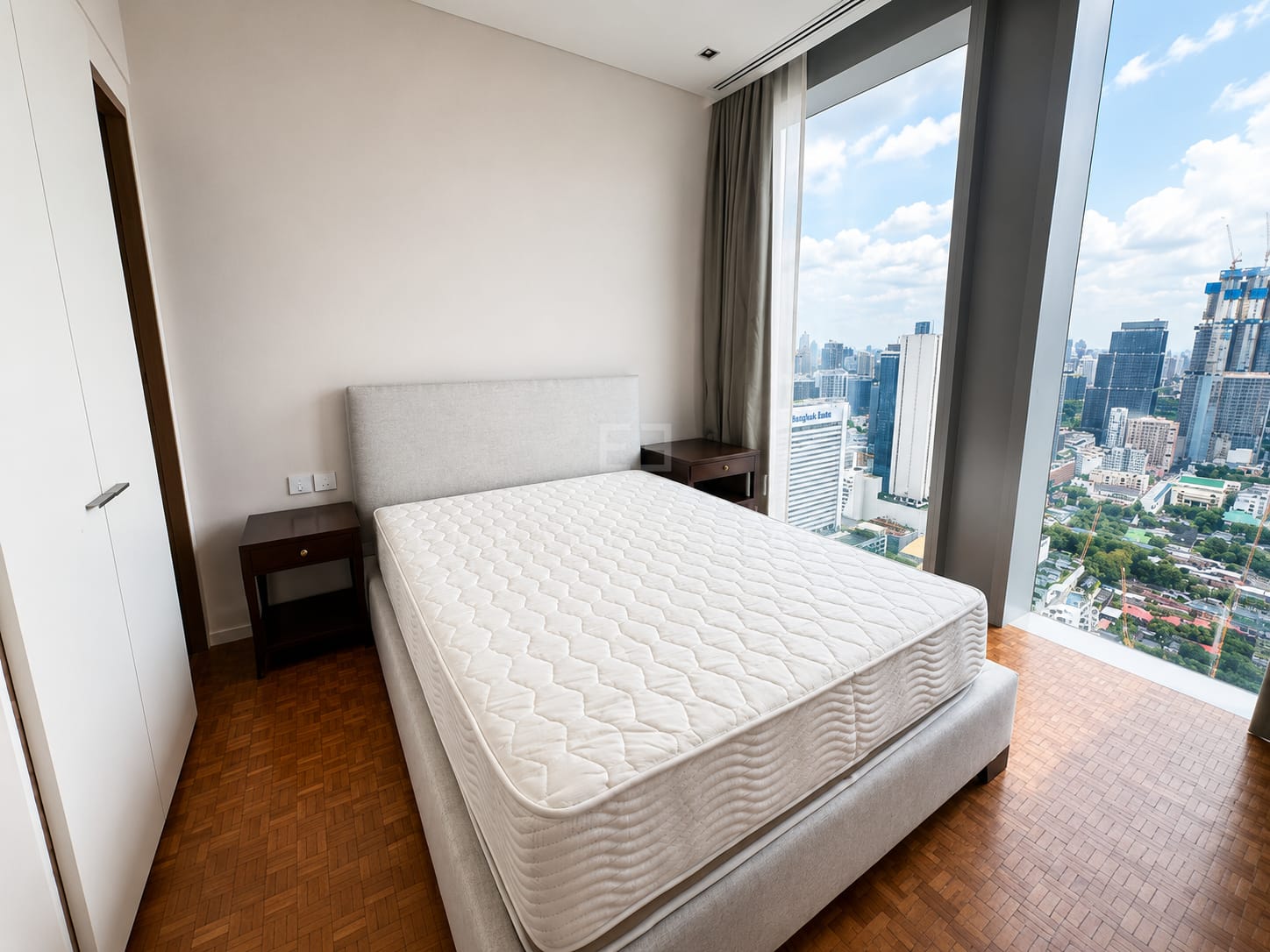 A bedroom at The Ritz-Carlton Residences Bangkok, a pale upholstered headboard beside corner glazing over central Bangkok