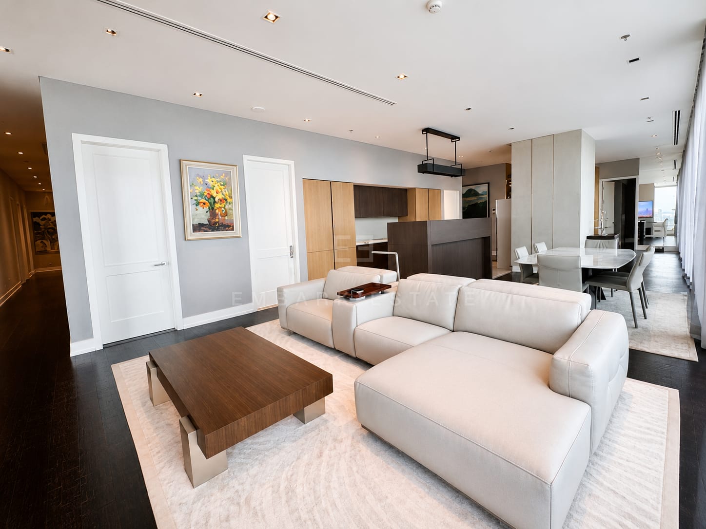 The living room at The Ritz-Carlton Residences Bangkok from the hall, the sectional and walnut table set on a rug with the kitchen and dining table beyond