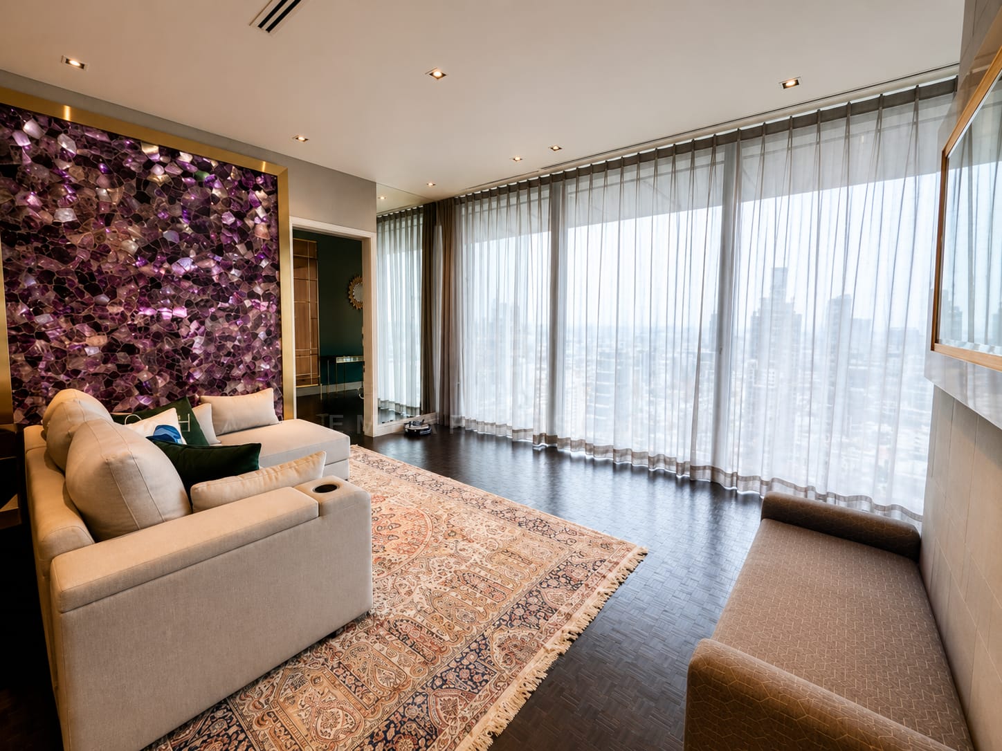 The sitting room at The Ritz-Carlton Residences Bangkok, a backlit amethyst stone wall above a sofa and Persian rug, with a window seat along the glazing