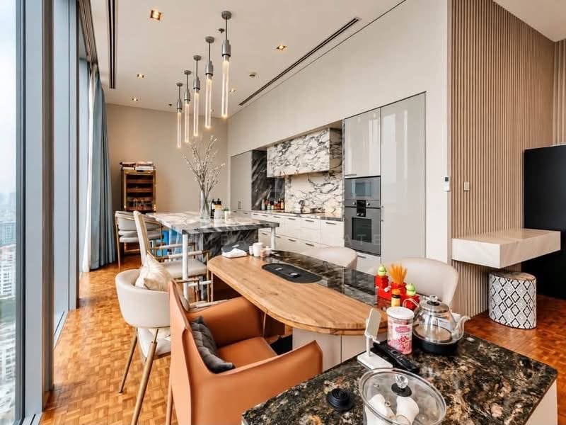 The kitchen at The Ritz-Carlton Residences Bangkok, a granite-topped island with bar seating, a marble splashback, built-in ovens and pendant lights, with the city at the window