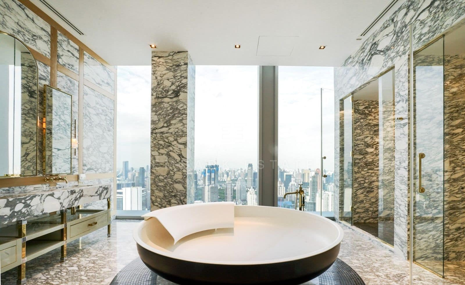 The master bathroom at The Ritz-Carlton Residences Bangkok, a round freestanding bath on a mosaic plinth, Arabescato marble walls trimmed in brass and floor-to-ceiling glass over the skyline