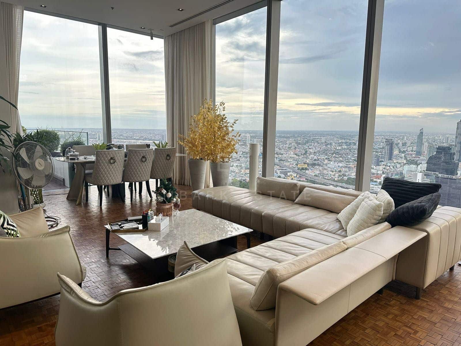The living room of the 400 square metre Sky Residence on level 61 at The Ritz-Carlton Residences Bangkok, two leather sofas facing across a marble table with the city along two walls of glass at dusk