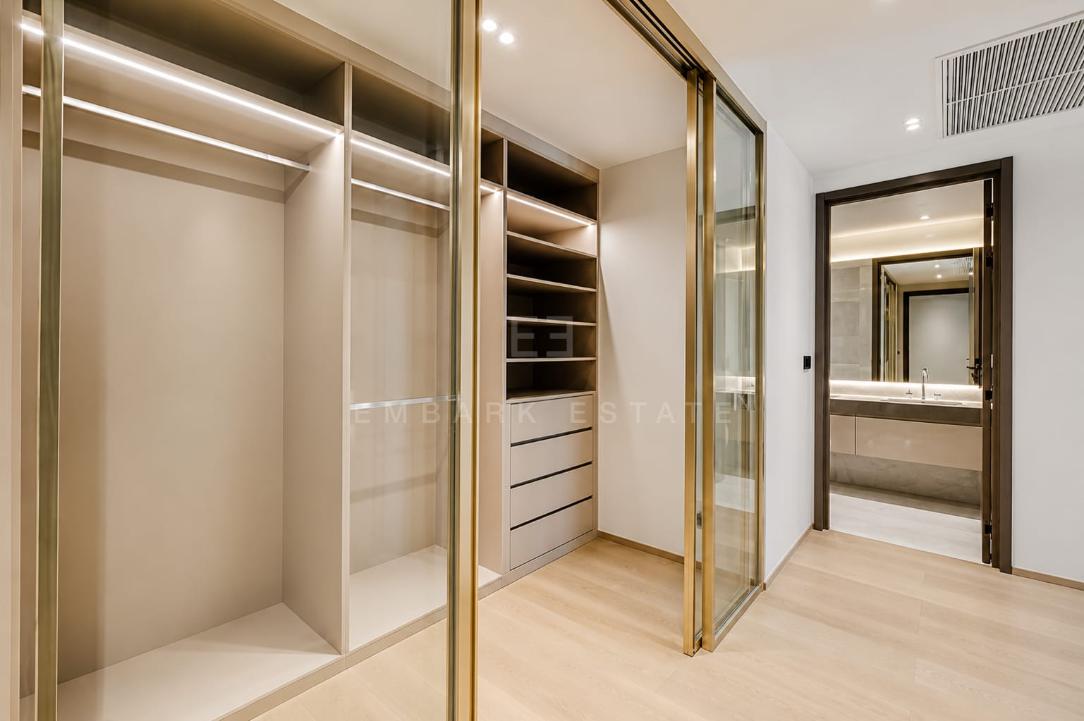 The walk-in wardrobe at Tonson One Residence, brass-framed glass doors onto fitted hanging and shelving with the bathroom lit beyond