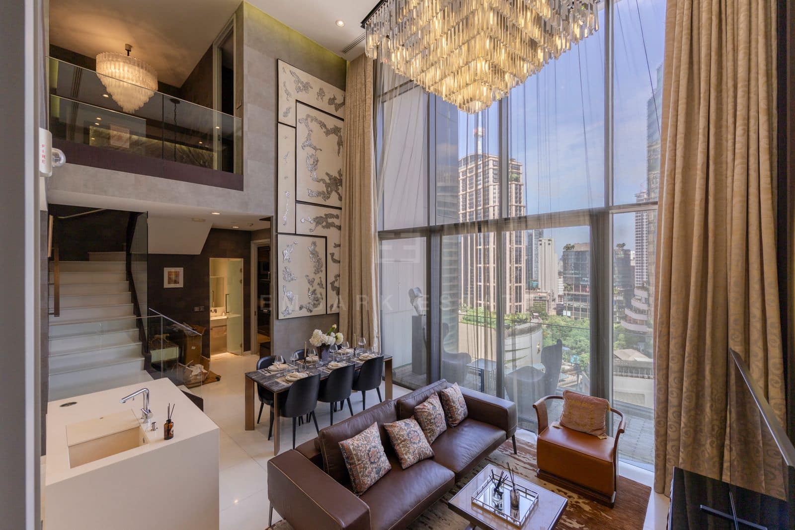 The double-height living room of the 168 square metre duplex at Vittorio Sukhumvit 39, a brown leather sofa and marble-topped dining table under a crystal chandelier, the staircase to the upper floor beyond