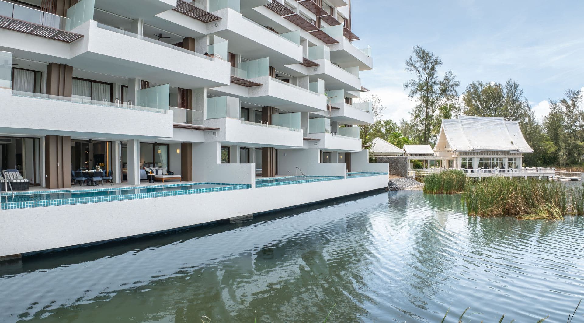 Photograph of Building 3 at Angsana Oceanview Residences, Laguna Phuket: the ground-floor homes' private pools along the lagoon edge, with a white Thai pavilion at the end of the building. Taken by the developer in October 2025