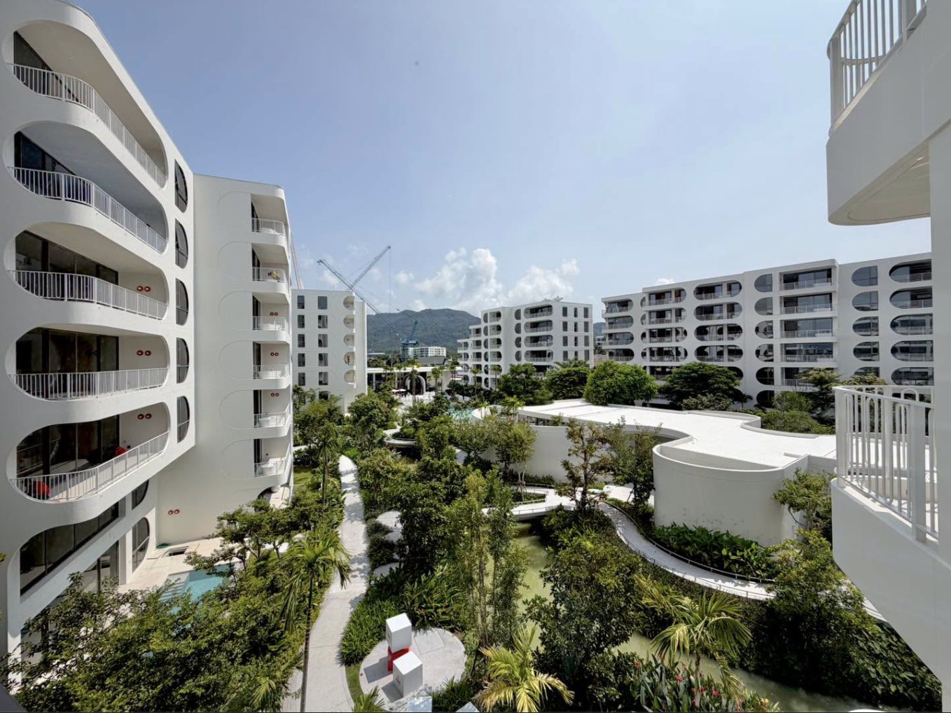 The finished white buildings of The Standard Residences, Phuket Bang Tao around their garden courtyard, with the curved balconies, the pool pavilion, the lagoon and the hills of Phuket behind, photographed at the opening in October 2026