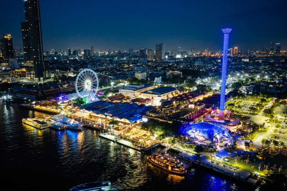 Asiatique The Riverfront at night from above, the Chao Phraya along the left, the ferris wheel and the lit observation tower behind, and the blue-lit Hatch Dome on the right of the site