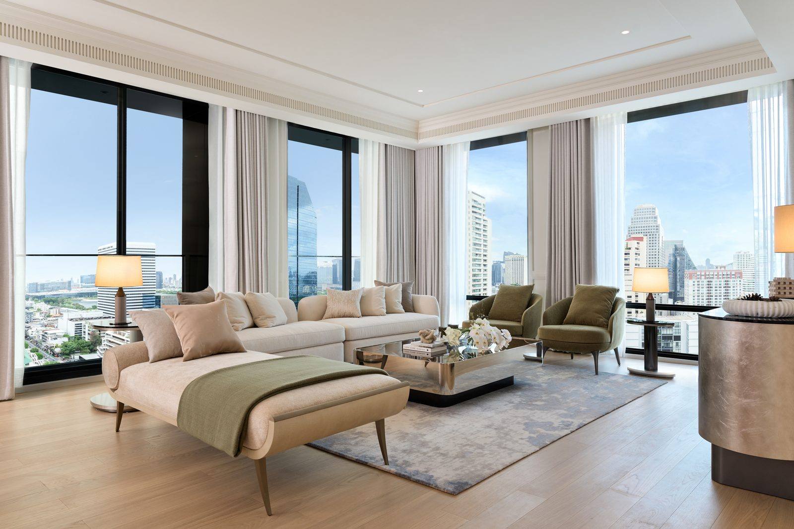 The living room of the show unit at InterContinental Residences Bangkok Asoke, photographed: a pale sofa and olive armchairs on a silk rug, a day bed at the window, and corner glazing onto the Bangkok skyline